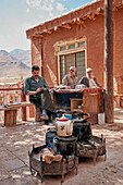 Menschen sitzen vor einem kleinen Teehaus, während ihr Tee auf traditionelle Weise über offenem Feuer zubereitet wird. Dorf Abyaneh, Kreis Natanz, Iran.