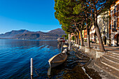 Altstadt mit Gebäuden und Bäumen am Ufer, Luganersee mit Boot an einem sonnigen Tag mit Bergen in Morcote, Tessin, Schweiz.