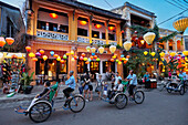 Cycle rickshaws with passengers drive by an old building with colorful lanterns illuminated at dusk. Hoi An Ancient Town, Quang Nam Province, Vietnam.