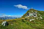  Woman sitting in front of Rifugio Mombarone hut with view of Colma di Mombarone peak with Christ statue, GTA, Grande Traversée des Alpes, Biella, Alpi Biellesi, Valais Alps, Piedmont, Italy 