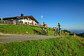  Man and woman mountain biking past alpine pasture, Gorialm, Kampenwand, Chiemgau Alps, Upper Bavaria, Bavaria, Germany 