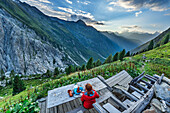  Woman hiking sits at a wooden table, takes a break and looks into the valley, Tour du Mont Blanc, Mont Blanc Group, Graian Alps, Valais, Switzerland 