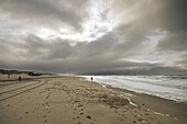 A cloudy beach scene with footprints in the sand and waves crashing on the shore.