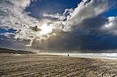 A dramatic beach scene with a cloudy sky and sunlight breaking through, casting shadows on the sand. The ocean waves are visible, and a few people are walking along the shore.