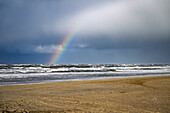 A serene beach scene with a rainbow arching over the ocean waves under a cloudy sky.