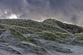 A dramatic landscape of sand dunes covered with grass under a stormy sky.