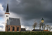 A church with a tall steeple stands against a dramatic cloudy sky. A vintage street lamp and a hedge are in the foreground, with bare trees visible in the background.