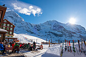 The Famous and Historical Hotel Bellevue des Alpes with people on the Terrace at 2061 Meters High Up and Mountain Peak of Mönch and Jungfrau and a Train in a Sunny Winter Day in Kleine Scheidegg, Grindelwald, Canton Bern, Switzerland.