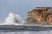  A large wave breaks on the cliffs below the Farol de Nazare lighthouse, Leiria, Portugal 