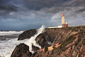  High waves crash against the cliffs near the Penedo da Saudade lighthouse. Cloudy sky, Marinha Grande, Leiria, Portugal 