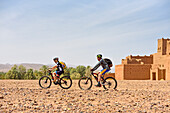Mountain bikers with electric assistance passing by a ruined ksar on the edge of a track through the reg, between Agdz and Zagora, Draa River valley, Province of Zagora, Region Draa-Tafilalet, Morocco, North West Africa