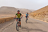 Mountain bikers with electric assistance on the road from Zagora to the south (N 9), Draa River valley, Province of Zagora, Region Draa-Tafilalet, Morocco, North West Africa