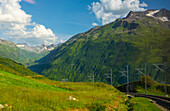 Railroad Tracks with View over a Mountain Valley and Blue Sky and Clouds in a Sunny Summer Day in Andermatt, Uri, Switzerland.