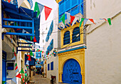  Alley with blue ornate wooden doors, Arabic balconies and windows, and colorful flags in the Medina of Hammamet, Tunisia 