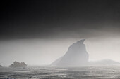  A foggy, silhouetted scene with passengers from an expedition cruise ship in a Zodiac rubber dinghy approaching an iceberg in the Vikingebugt, Arctic Greenland, Europe 