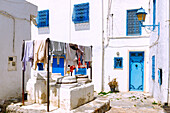 Gasse mit Wäsche zum Trocknen über antiken Säulenbasen und weiße Kubenhäuser mit blauen Holztüren und Fenstergittern in der (Medina) Altstadt von Sidi Bou Said, Tunesien