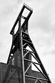 A black and white photograph of a large industrial structure, featuring a towering headframe with a triangular design. The building has a modern architectural style, showcasing steel and glass elements against a cloudy sky.