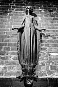 A black and white photograph of a statue depicting a figure with outstretched arms, set against a textured brick wall. The statue has intricate details and a serene expression, symbolizing grace and spirituality.