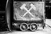 A vintage mining cart with a weathered surface, featuring two crossed hammers and the word 'Glück Auf' painted in white. The scene is captured in black and white, emphasizing the industrial atmosphere.