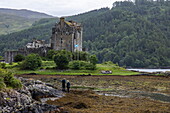  Two people standing on rocks near Eilean Donan Castle (filming location of &quot;Highlander&quot;) at the meeting point of Loch Duich, Loch Long and Loch Alsh, Kyle Dornie, Highland Council, Scotland, United Kingdom, Europe 