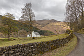  A small white Scottish cottage stands on a small street behind a natural stone wall. 