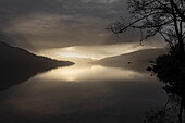 Goldene Morgenstimmung mit einer Wolkenlücke am Ufer See Loch Katrine. Die Wolken und Berge spiegeln sich im Wasser, Schottland, Großbritannien
