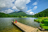  Man and woman sitting on a wooden jetty in Lake Spitzingsee, Brecherspitz mountain in the background, Spitzing, Mangfall Mountains, Bavarian Alps, Upper Bavaria, Bavaria, Germany 