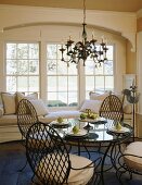 Metallstühle um runden Esstisch mit Glasplatte und Polsterbank vor der Fensterfront in elegantem Landhaus-Wohnzimmer
