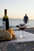 Red wine and bread on table by sea, person in background
