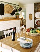 Country house kitchen with rustic dining table, stack of plates, wine glasses, red wine and basket of vegetables