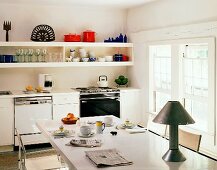 Simple, cheerful kitchen with crockery on long shelves and table lamp on white table