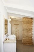 Ein Muschelvorhang und ein Raumtrenner aus Holzstäben im Badezimmer mit dezentem Waschtisch
