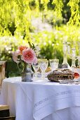 Bread, wine glasses and jug of roses on laid table in garden