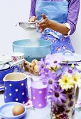 Woman sieving flour into a bowl, crockery & eggs in front