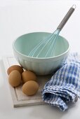 Mixing bowl with whisk, eggs, tea towel on chopping board