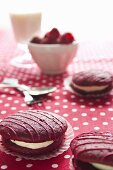 Raspberry whoopie pies on a spotted tablecloth with fresh raspberries, a glass of milk and cutlery