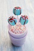 Blue gift-shaped cake pops with red sugar bows in a cup of colourful sugar strands