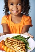 A young girl holding a plate of grilled chicken, grilled peaches and beans