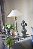 An antique table lamp with a white shade between a flower vase and an angel statue