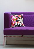 A purple sofa with arm and back rests and a colourful cushion in the corner