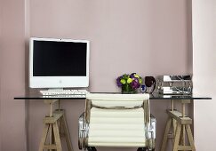 Arbeitsplatz vor pastellfarbener Wand mit Schreibtisch aus Glasplatte & Holzböcken, Computer & Leder-Bürostuhl