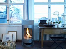 Stainless steel wood burning stove with fire next to a wooden table in front of a wall of windows