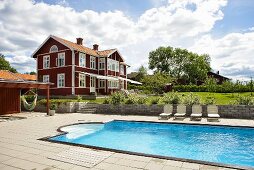 Loungers by a pool and a red-brown wooden house with white windows in the background