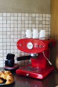 A red espresso machine with coffee cups and a croissant on a plate in the corner of a kitchen with white mosaic tiles