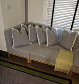 Hellgraues Sofa und Kissen in gleichem Bezug in Fensternische vor geschlossener Jalousie