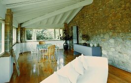 White sofa and dining area under a beam ceiling and natural stone wall in the living room of a country home