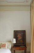 Corner of a bedroom -- a painting on a nightstand with a table lamp with a white shade