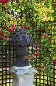 Blumentopf aus Stein auf Stele vor rosenberanktem Gerüst