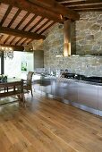 Stainless steel kitchen in front of a natural stone wall and beam ceiling in a renovated country home