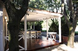 Trees in front of white wooden terrace with posts and a patio table on a raised wooden platform